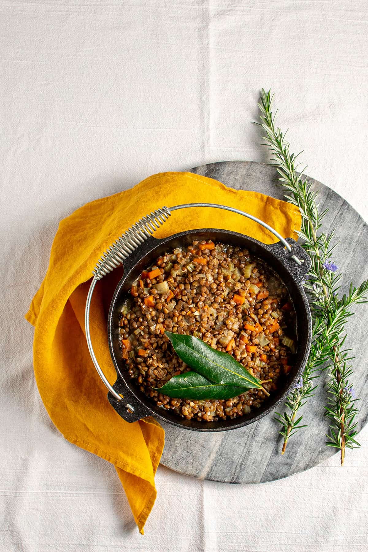 A cast iron pot of lentils, sitting on a marble chopping board with an orange linen napkin alongside. Inside the pot there are cooked lentils with small chunks of celery and carrot, and two bay leaves sitting on top. 