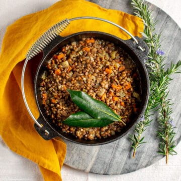 A cast iron pot of lentils, sitting on a marble chopping board with an orange linen napkin alongside. Inside the pot there are cooked lentils with small chunks of celery and carrot, and two bay leaves sitting on top.