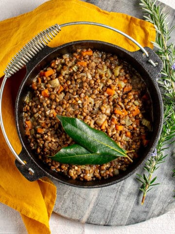 A cast iron pot of lentils, sitting on a marble chopping board with an orange linen napkin alongside. Inside the pot there are cooked lentils with small chunks of celery and carrot, and two bay leaves sitting on top.