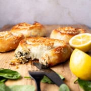 A front on view of baked filo pastry borek, rolled in small spiral shapes. A scattering of spinach leaves are nearby, and two cut lemon halves. One of the borek is partly eaten and you can see the creamy filling.