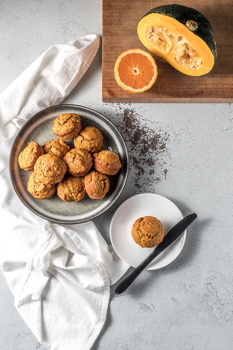 Pumpkin, orange and chia seed muffins on a counter top, pictured alongside a cut pumpkin and half an orange. Vegan. 