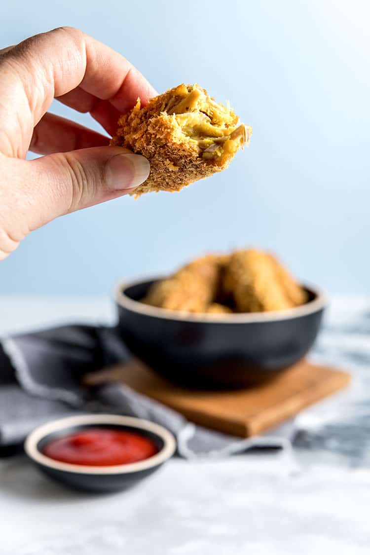 A hand holding a jackfruit and chickpea nugget, with a bite taken from it, showing the moist and textured interior of the nugget. There is a bowl of vegan nuggets and tomato ketchup in the background. 