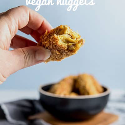 A hand holding a jackfruit and chickpea nugget, with a bite taken from it, showing the moist and textured interior of the nugget. There is a bowl of vegan nuggets and tomato ketchup in the background.