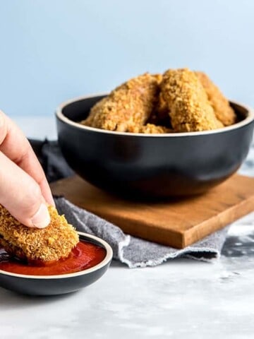 Jackfruit and chickpea vegan nuggets in a black bowl, with a person's hand holding a nugget and dipping it into tomato ketchup.