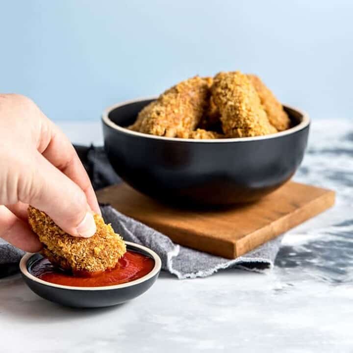 Jackfruit and chickpea vegan nuggets in a black bowl, with a person's hand holding a nugget and dipping it into tomato ketchup.