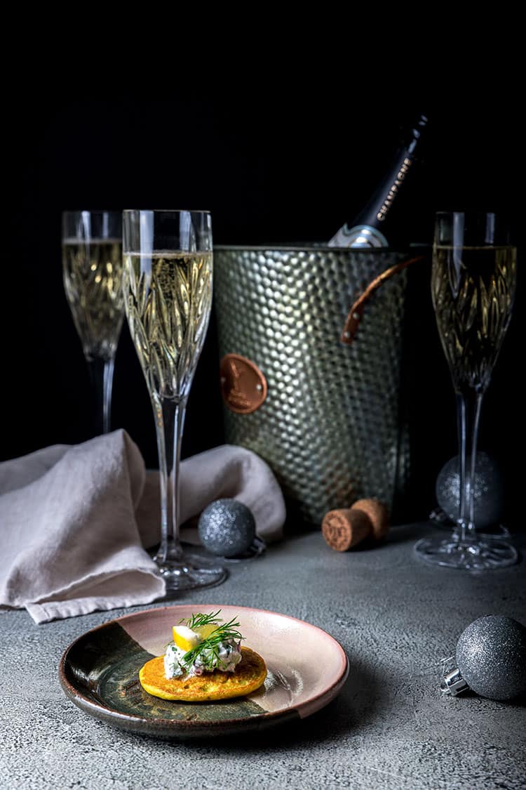 A vegan blini on a small side plate, with champagne flutes, a champagne bucket and bottle of champagne in the background.