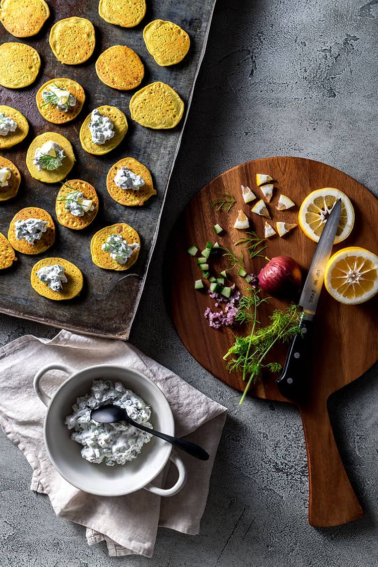 A tray full of spiced blini, alongside a board with chopped lemon, fennel fronds, cucumber and red onion. 