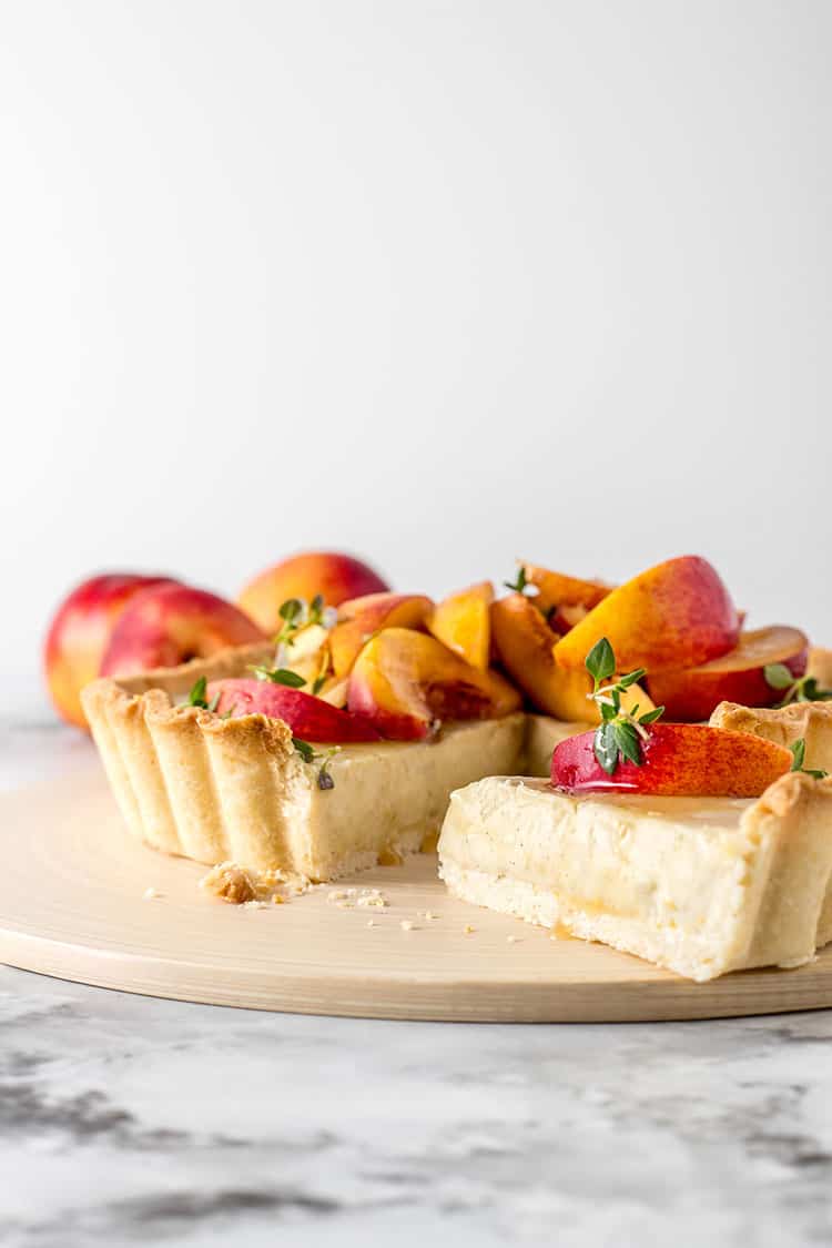 A vegan custard tart sitting on a chopping board, with a slice removed. The tart is topped with sliced nectarines, drizzled with honey and sprinkled with fresh thyme leaves. 