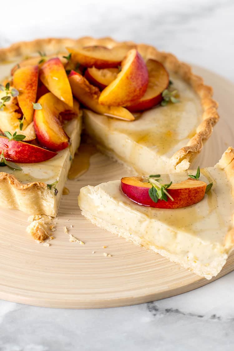 A vegan custard tart sitting on a chopping board, with a slice removed. The tart is topped with sliced nectarines, drizzled with honey and sprinkled with fresh thyme leaves.