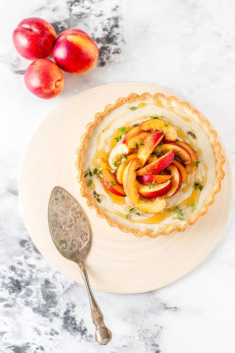 A vegan custard tart sitting on a chopping board. The tart is topped with sliced nectarines, drizzled with honey and sprinkled with fresh thyme leaves.