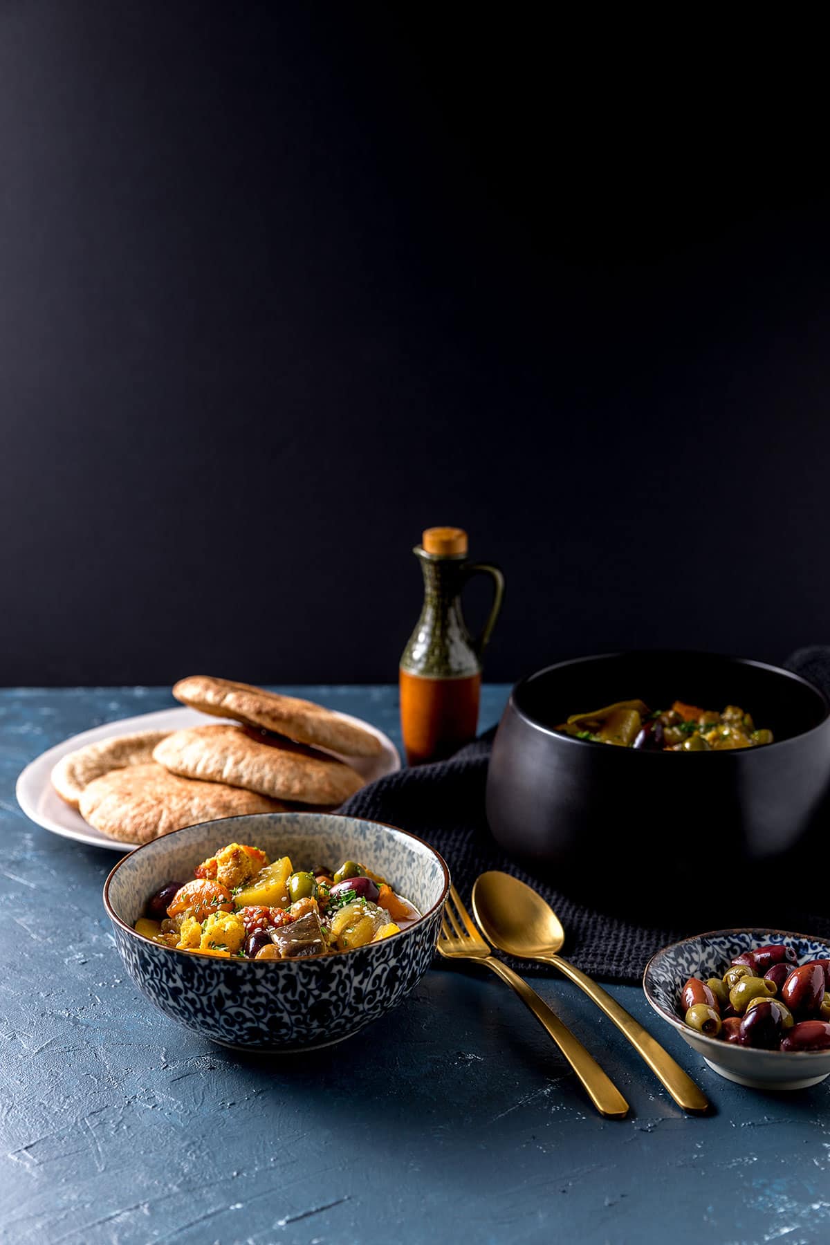 A serving bowl full of golden coloured vegetable and olive tagine, which is vegan and gluten free. Some has been served into a smaller bowl, sitting alongside a plate of flat breads. 