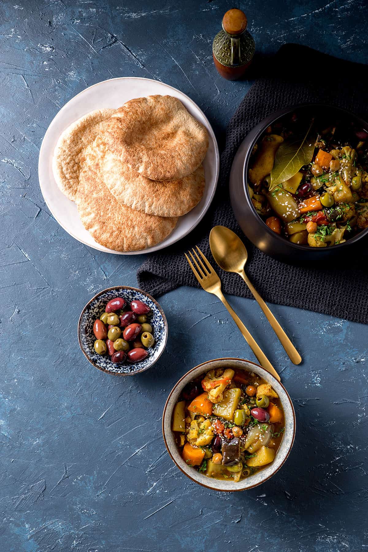 A serving bowl full of golden coloured vegetable and olive tagine, which is vegan and gluten free. Some has been served into a smaller bowl, sitting alongside a plate of flat breads.