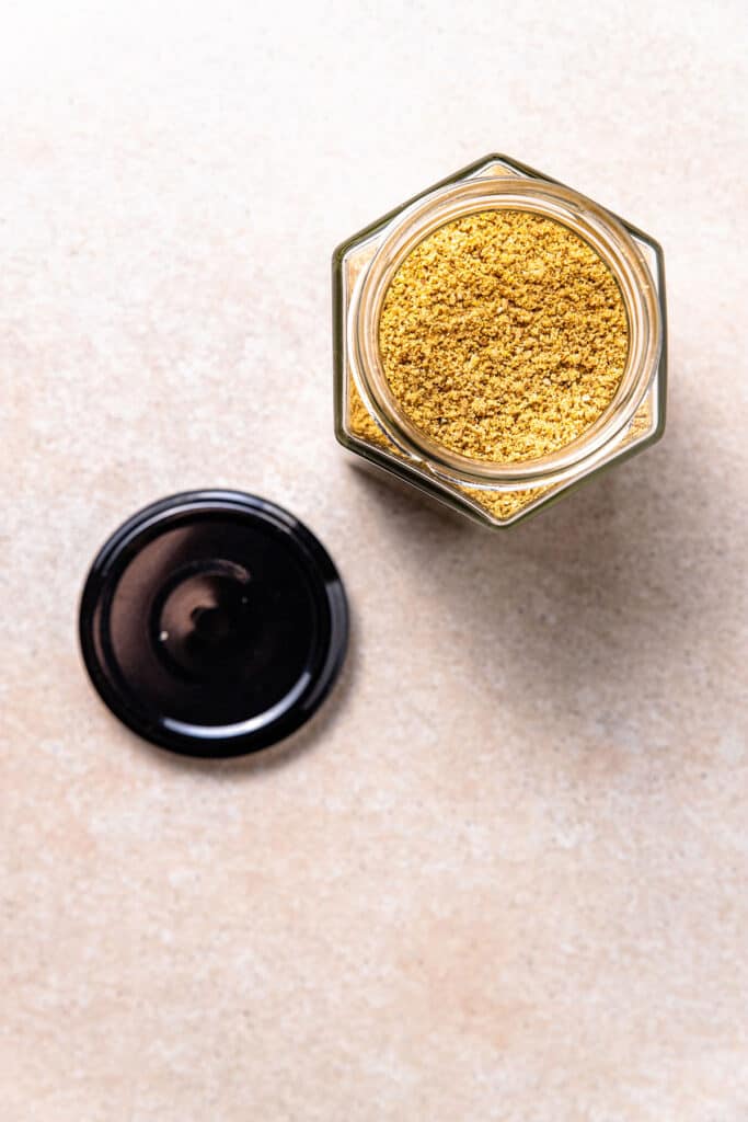 A hexagonal shaped jar full of golden coloured vegan parmesan sprinkle. The jar is open with the lid sitting beside it. 