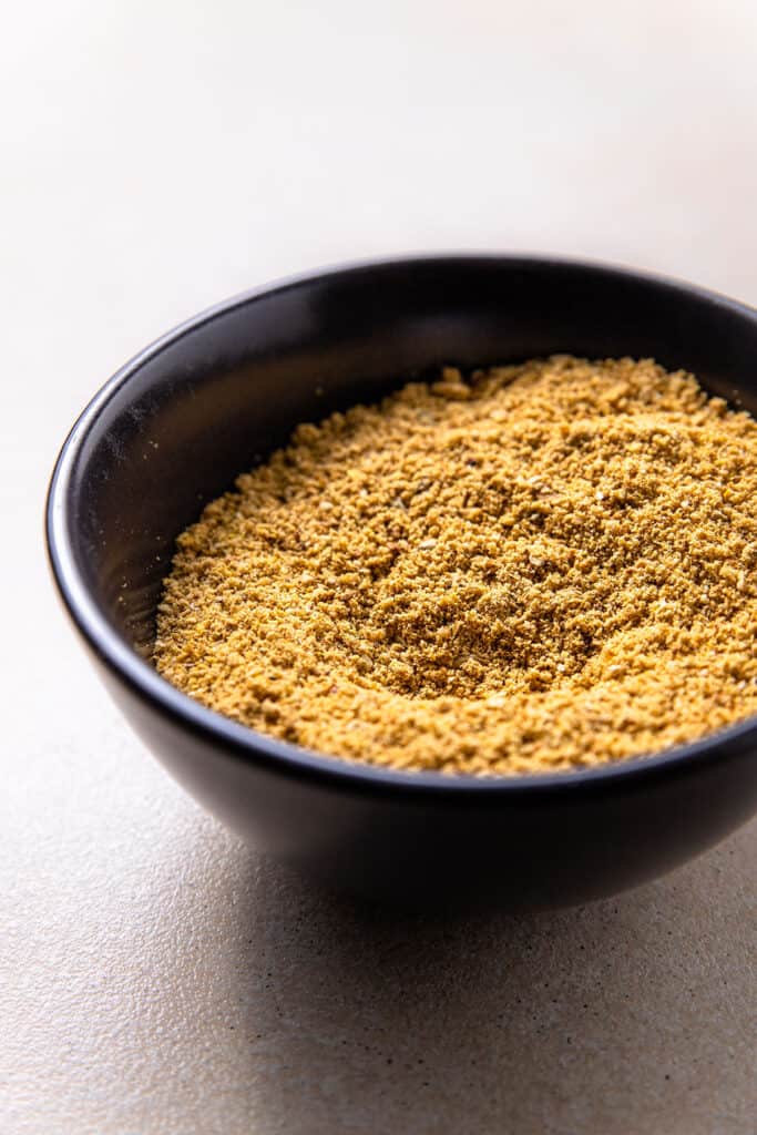 A black bowl holding vegan parmesan sprinkle. The texture is a coarse powder, and the colour is golden brown. 