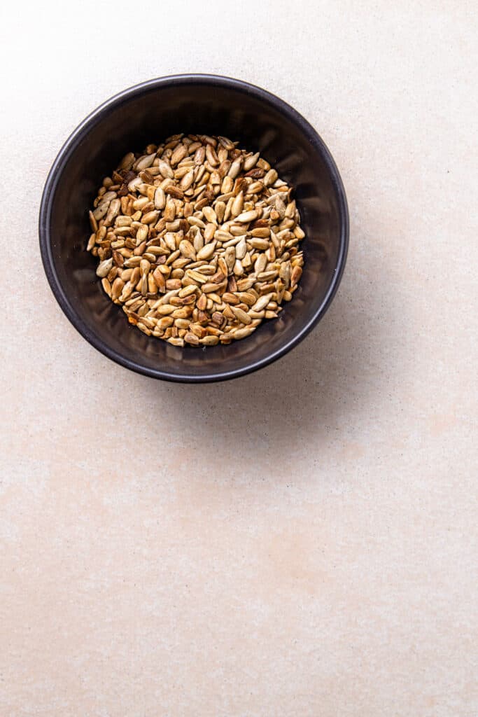 A black bowl holding golden brown, toasted sunflower seeds. The bowl is sitting on a beige counter top. 