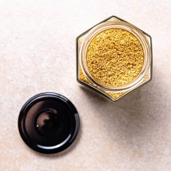A hexagonal shaped jar full of golden coloured vegan parmesan sprinkle. The jar is open with the lid sitting beside it.