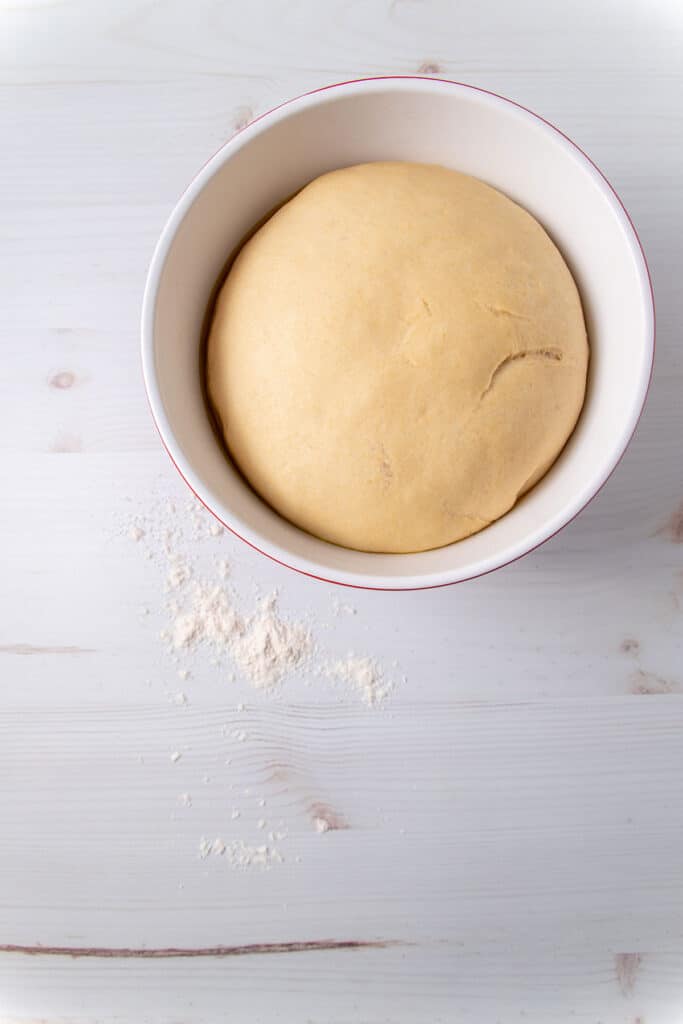 A mixing bowl sitting on a floured benchtop, with a ball of pizza dough inside. The pizza dough has risen and has a smooth surface. 