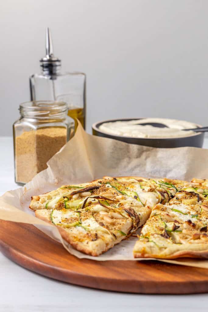 Sliced pizza sitting on baking paper on a chopping board, with a bottle of olive oil, jar of vegan parmesan sprinkle and bowl of white sauce in the background. 