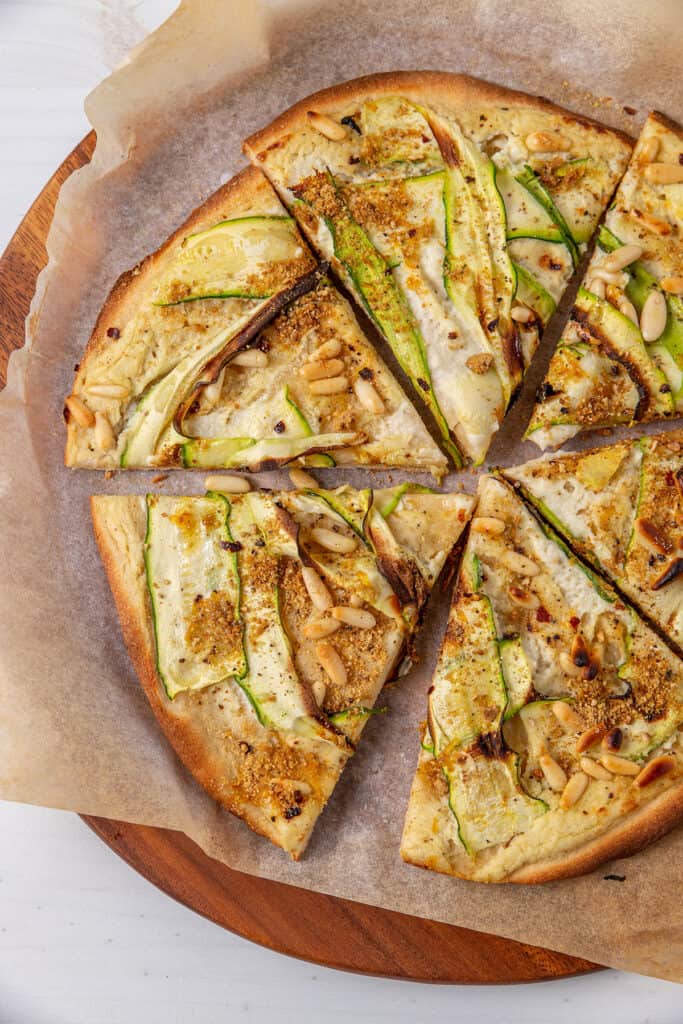 A close up view of a pizza cut into six pieces sitting on a chopping board. The pizza is topped with white sauce, zucchini ribbons, pine nuts and vegan parmesan sprinkle.