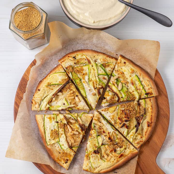 A pizza cut into six pieces sitting on a chopping board. The pizza is topped with white sauce, zucchini ribbons, pine nuts and vegan parmesan sprinkle.