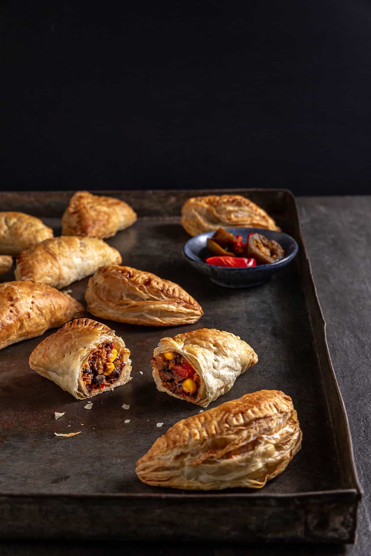 A metal tray with cooked hand pies on it. One is cut in half to reveal the black bean based filling. 