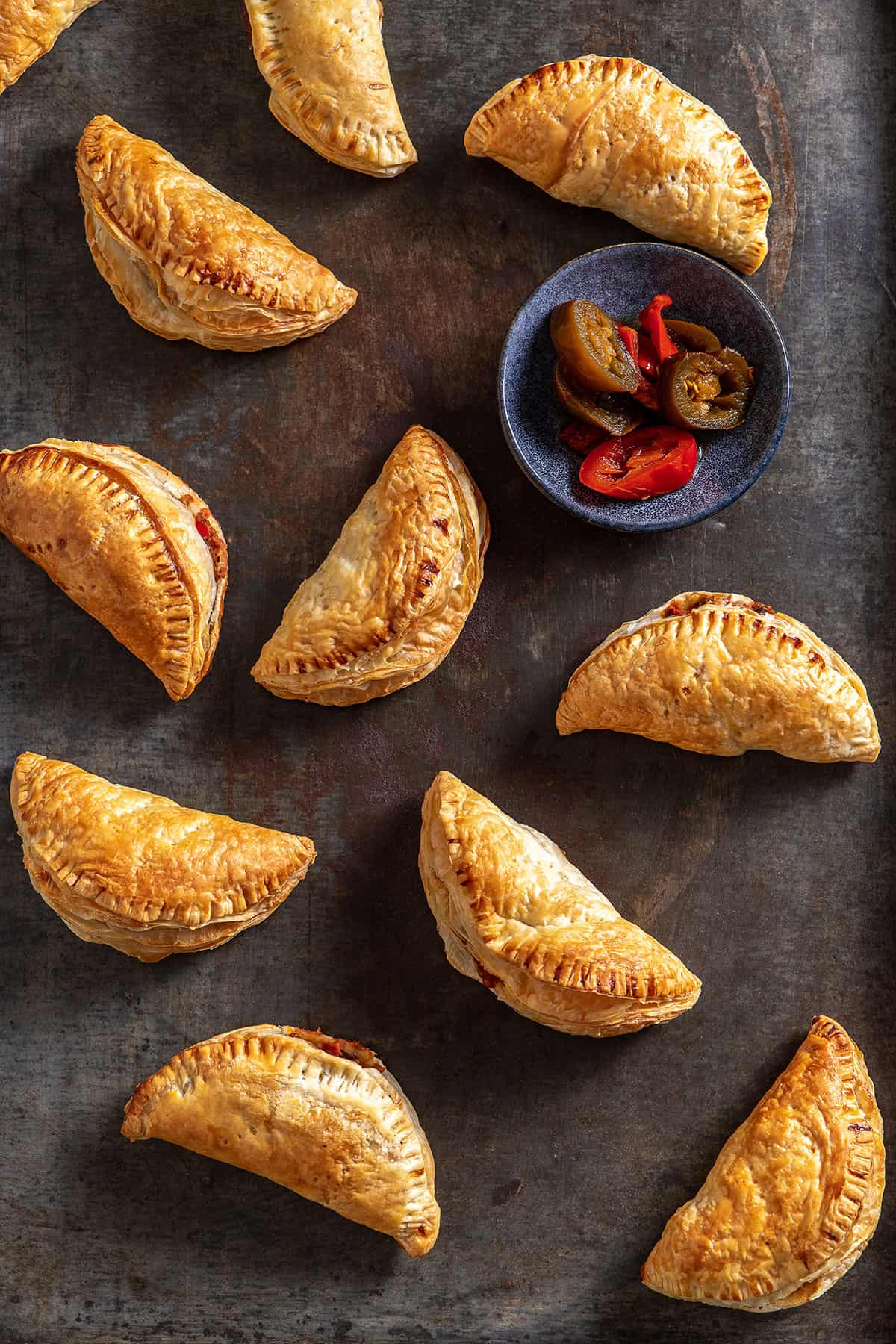 A metal tray with cooked hand pies on it, and a small dish of pickled jalapenos.