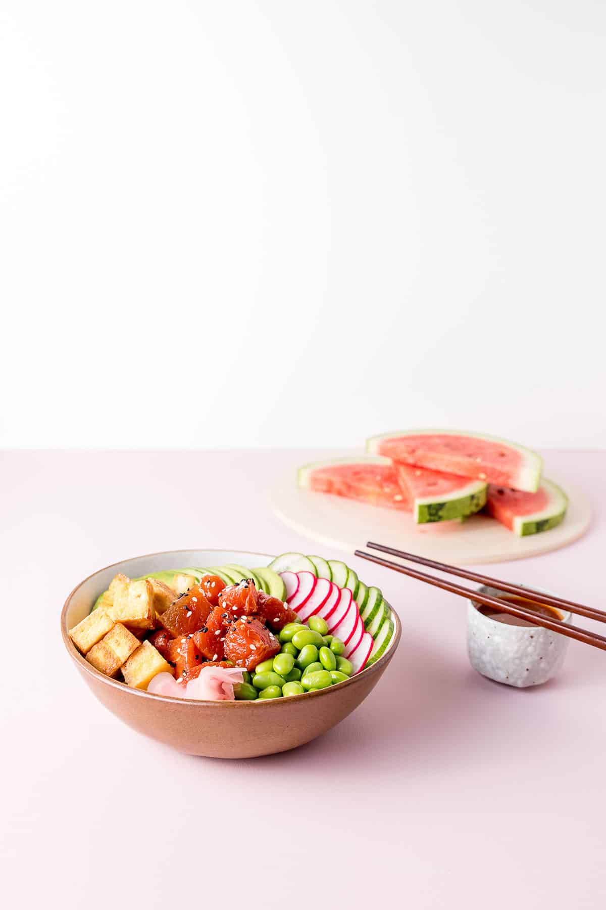 Picture of a watermelon poke bowl (A bowl with edamame beans, sliced radish, cucumber and avocado, fried tofu and marinated watermelon visible), sliced watermelon on a chopping board, and a small dish of watermelon poke marinade.