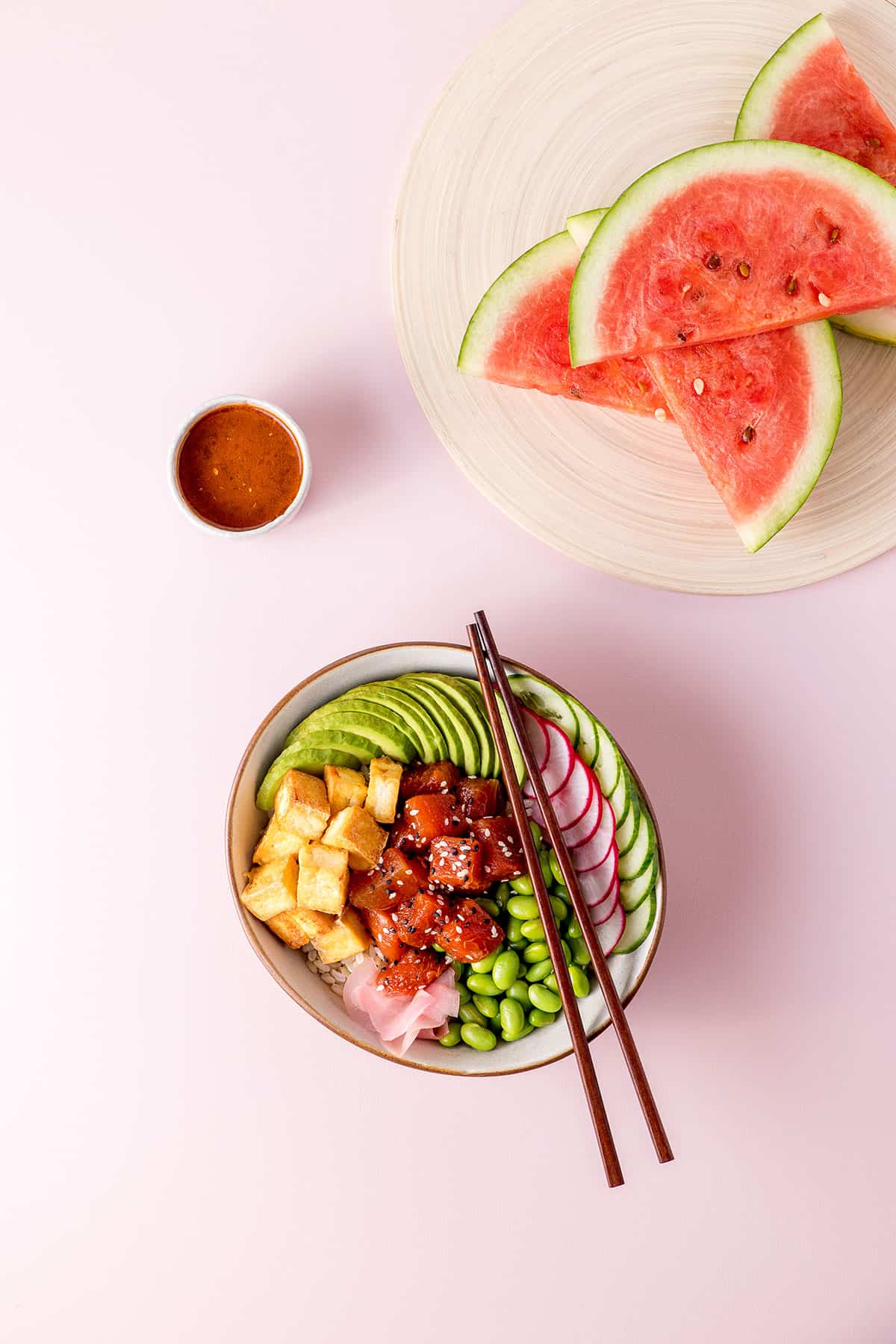 Overhead shot of a watermelon poke bowl (a bowl with edamame beans, sliced radish, cucumber and avocado, fried tofu and marinated watermelon visible), sliced watermelon on a chopping board, and a small dish of watermelon poke marinade. 
