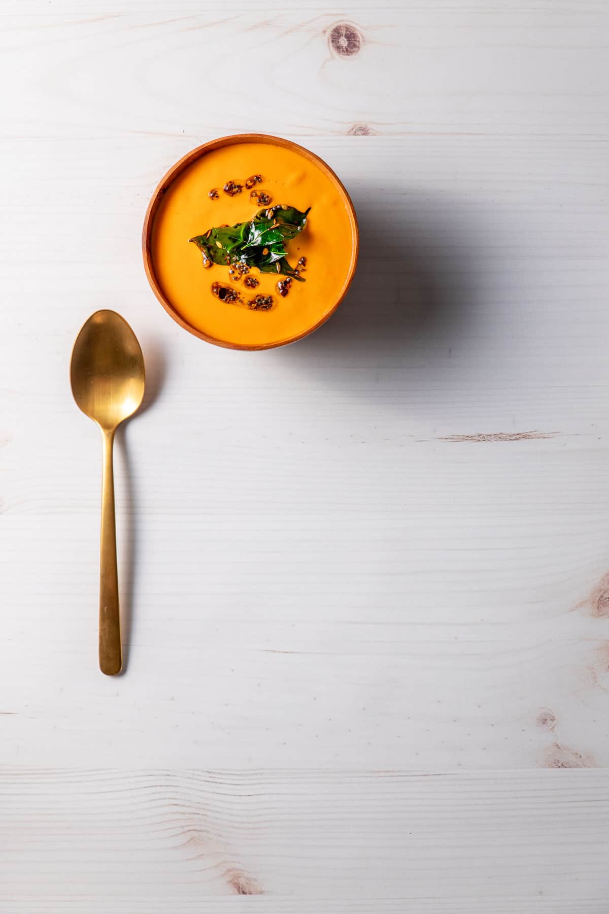 A wooden bowl full of bright orange-red creamy tomato and cashew soup, topped with oil, whole spices and fried curry leaves. A brass spoon sits next to the bowl.