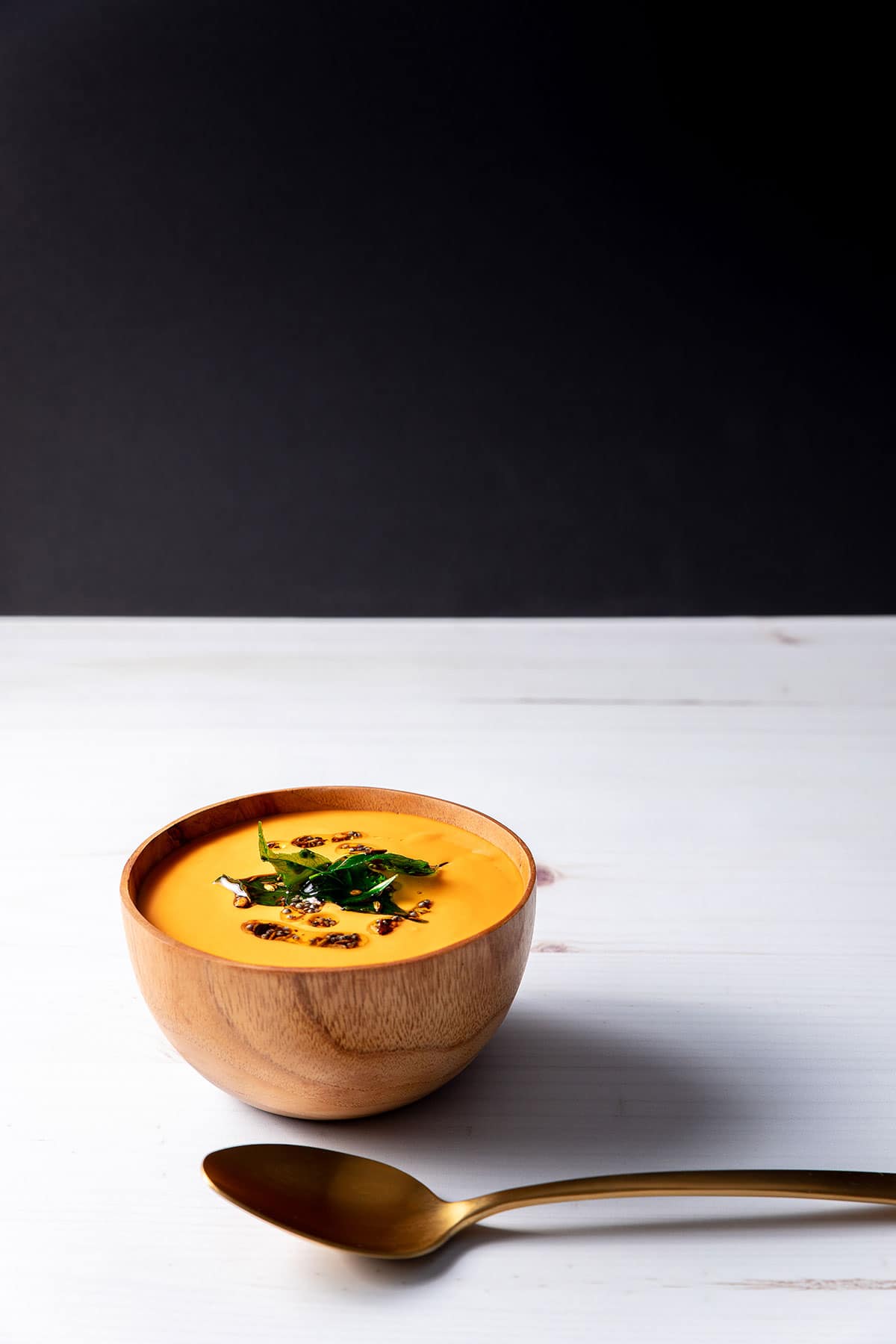 A wooden bowl full of bright orange-red creamy tomato and cashew soup, topped with oil, whole spices and fried curry leaves. A brass spoon sits next to the bowl.