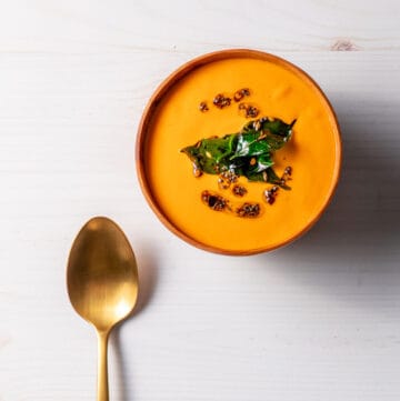 A wooden bowl full of bright orange-red creamy tomato and cashew soup, topped with oil, whole spices and fried curry leaves. A brass spoon sits next to the bowl.
