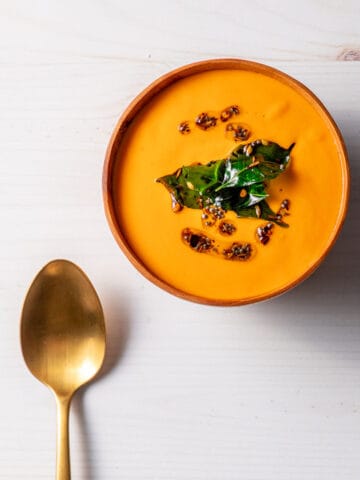 A wooden bowl full of bright orange-red creamy tomato and cashew soup, topped with oil, whole spices and fried curry leaves. A brass spoon sits next to the bowl.