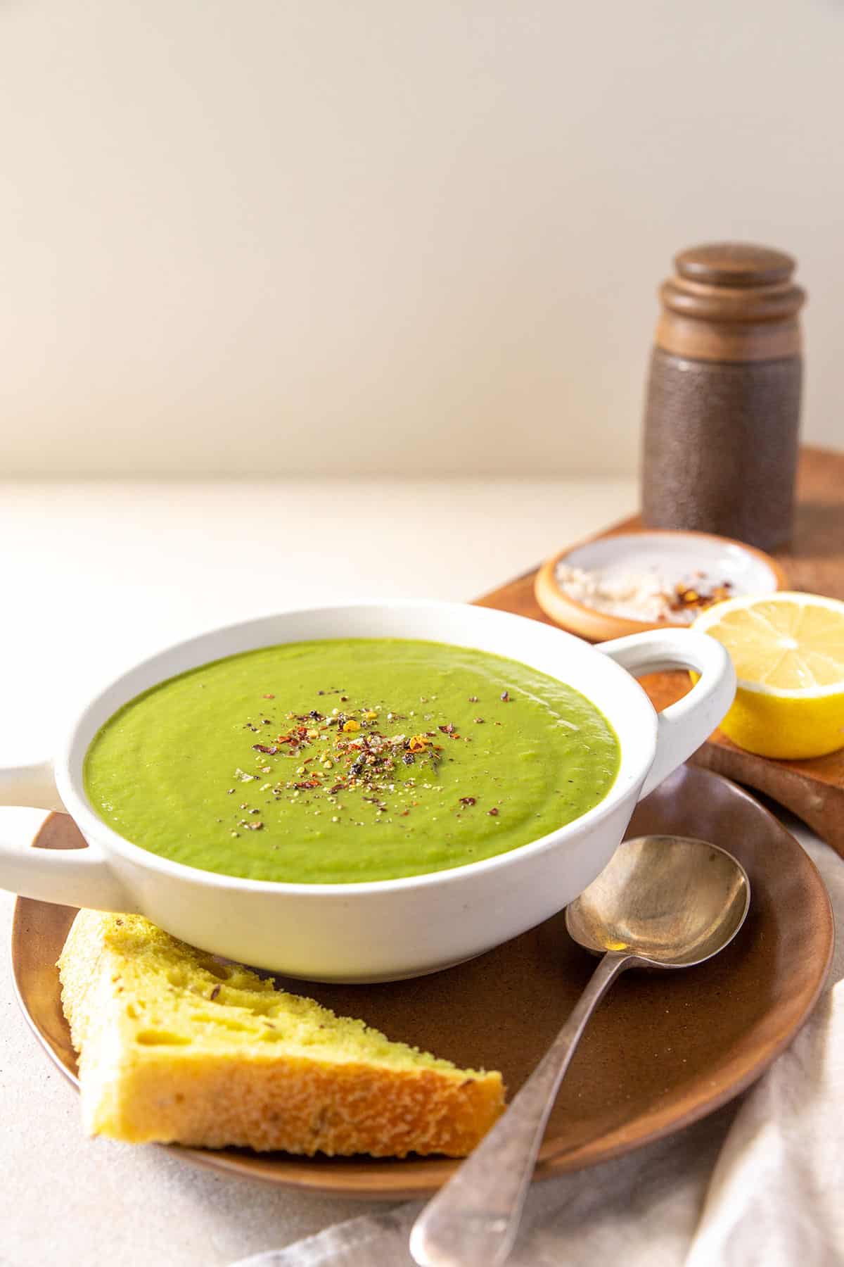 A white bowl of bright green broccoli soup, sitting on a brown plate with a slice of bread beside it. In the background is a board with half a lemon, a dish of salt and chilli flakes, and a pepper shaker. 