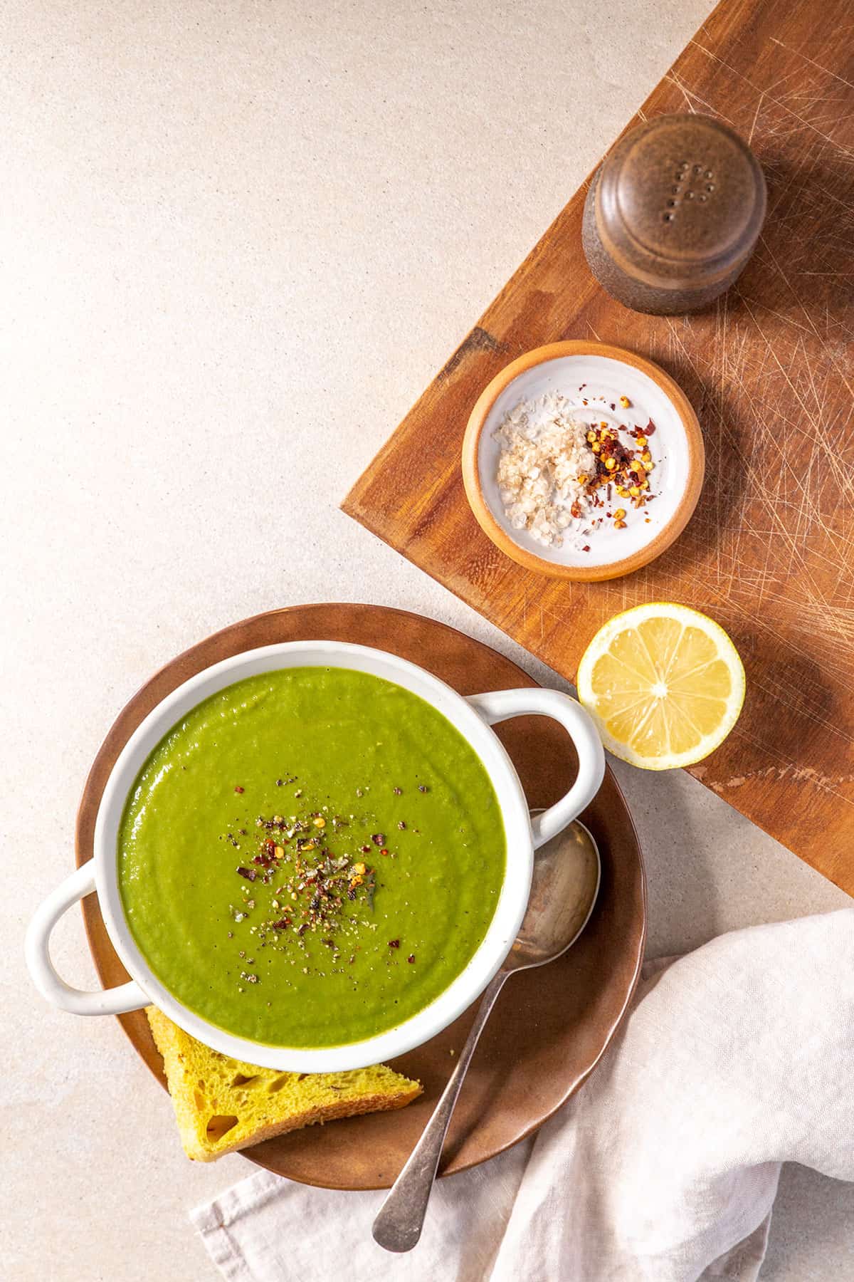 A bowl of green broccoli soup, sitting on a small plate with a slice of bread. Also pictured: salt, pepper, chilli flakes and a lemon for squeezing. 