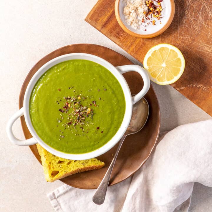 A bowl of green broccoli soup, sitting on a small plate with a slice of bread. Also pictured: salt, pepper, chilli flakes and a lemon for squeezing.