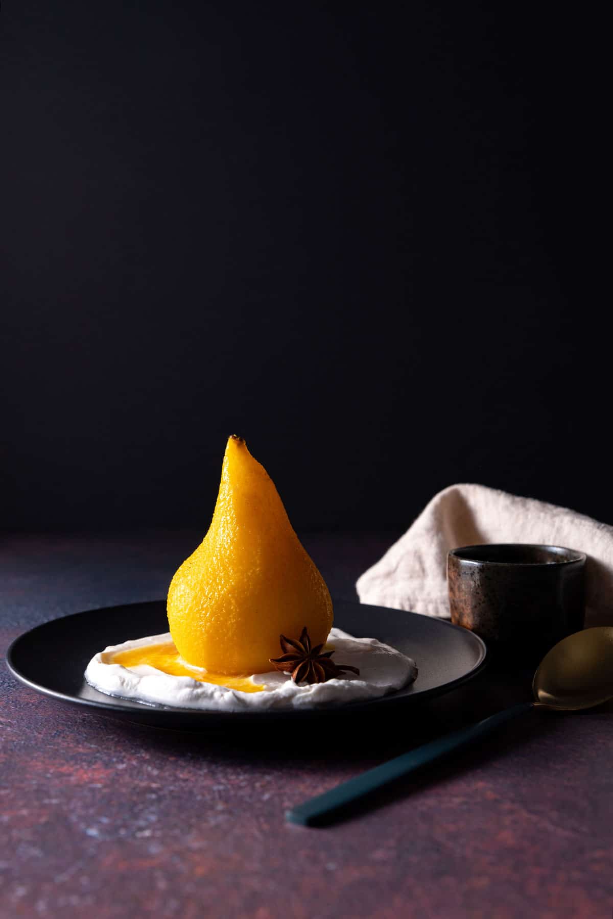 A whole, golden coloured poached pear sitting on a black plate, with coconut yoghurt and a star anise. 