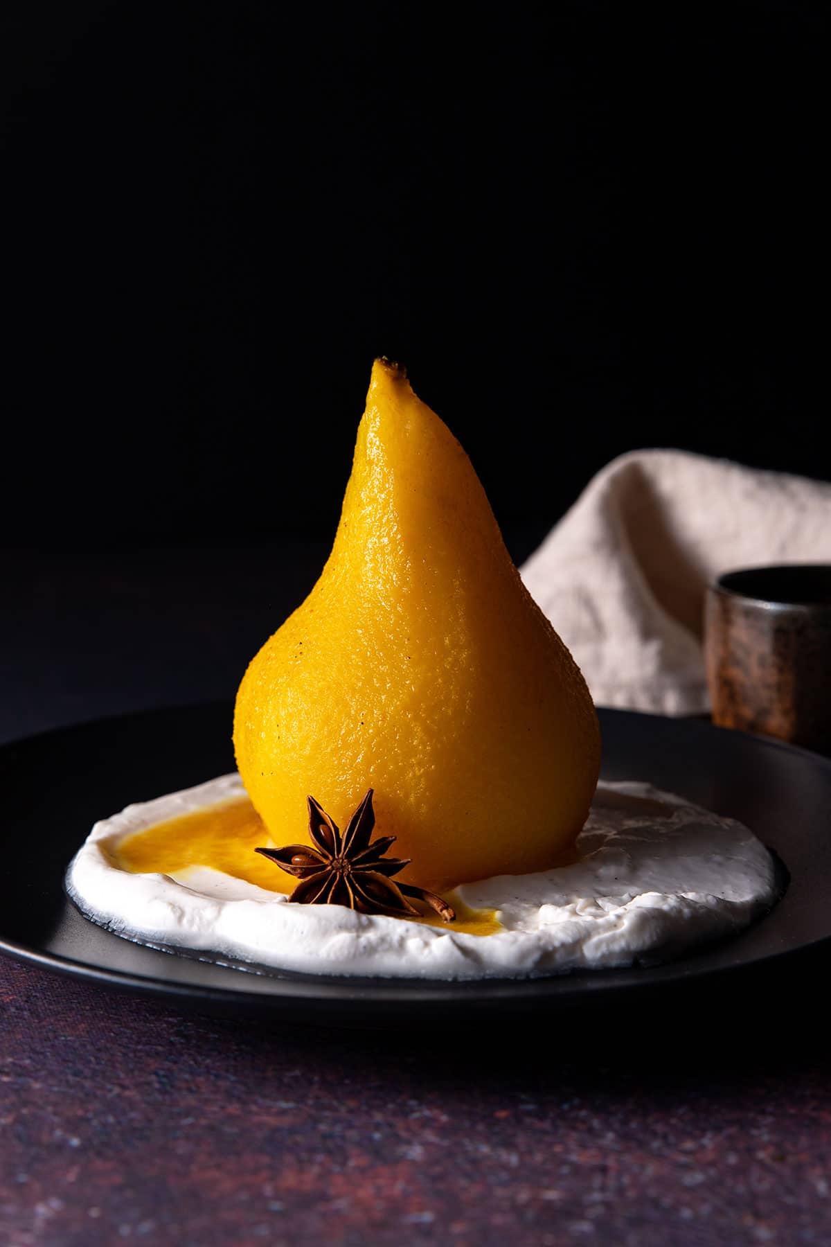 A close up picture of a whole, golden coloured poached pear sitting on a black plate, with coconut yoghurt, a drizzle of poaching liquid, and a star anise.