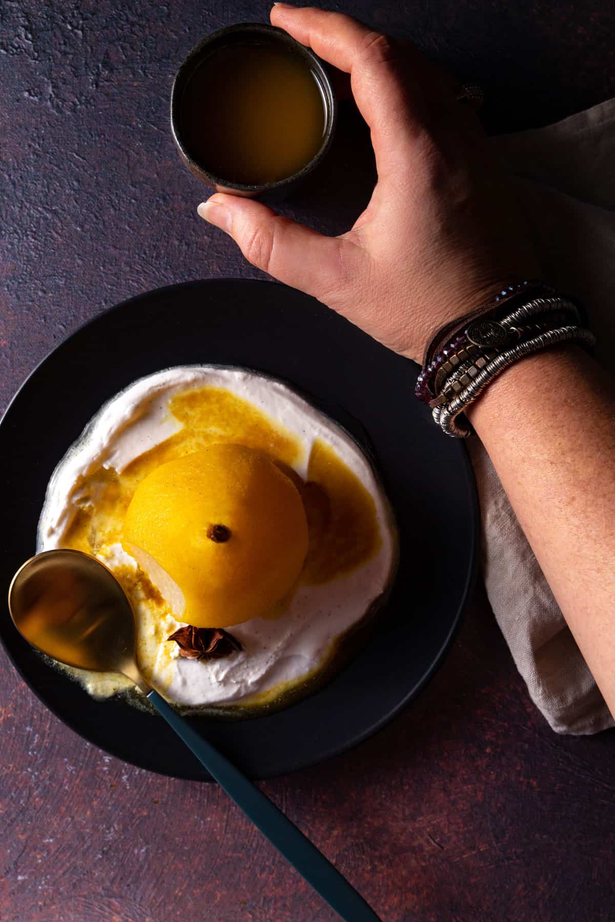 An overhead photo of a golden poached pear on a black plate, with coconut yoghurt, a star anise, a puddle of poaching liquid, and a hand picking up a small container of extra poaching liquid for pouring. 