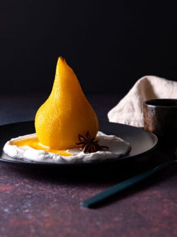 A whole, golden coloured poached pear sitting on a black plate, with coconut yoghurt and a star anise.