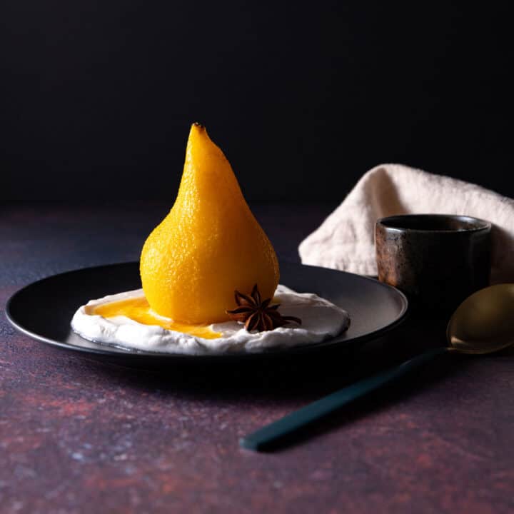 A whole, golden coloured poached pear sitting on a black plate, with coconut yoghurt and a star anise.