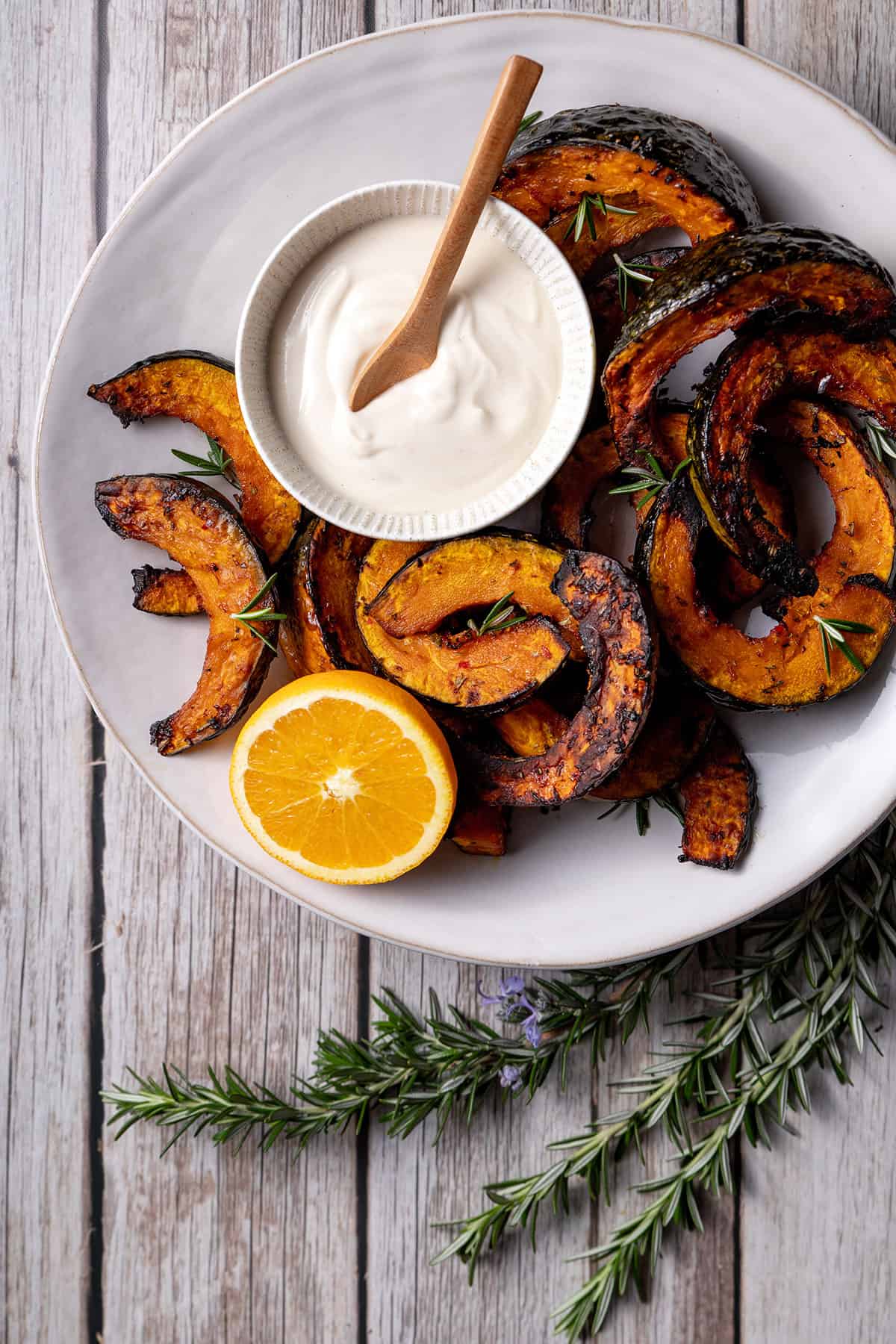 A white plate with slices of roast buttercup pumpkin, pictured with a small bowl of coconut yoghurt and tahini sauce, half an orange and a sprig of rosemary. 