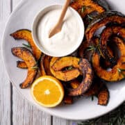 A white plate with slices of roast buttercup pumpkin, pictured with a small bowl of coconut yoghurt and tahini sauce, half an orange and some small sprigs of rosemary.