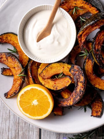 A white plate with slices of roast buttercup pumpkin, pictured with a small bowl of coconut yoghurt and tahini sauce, half an orange and some small sprigs of rosemary.
