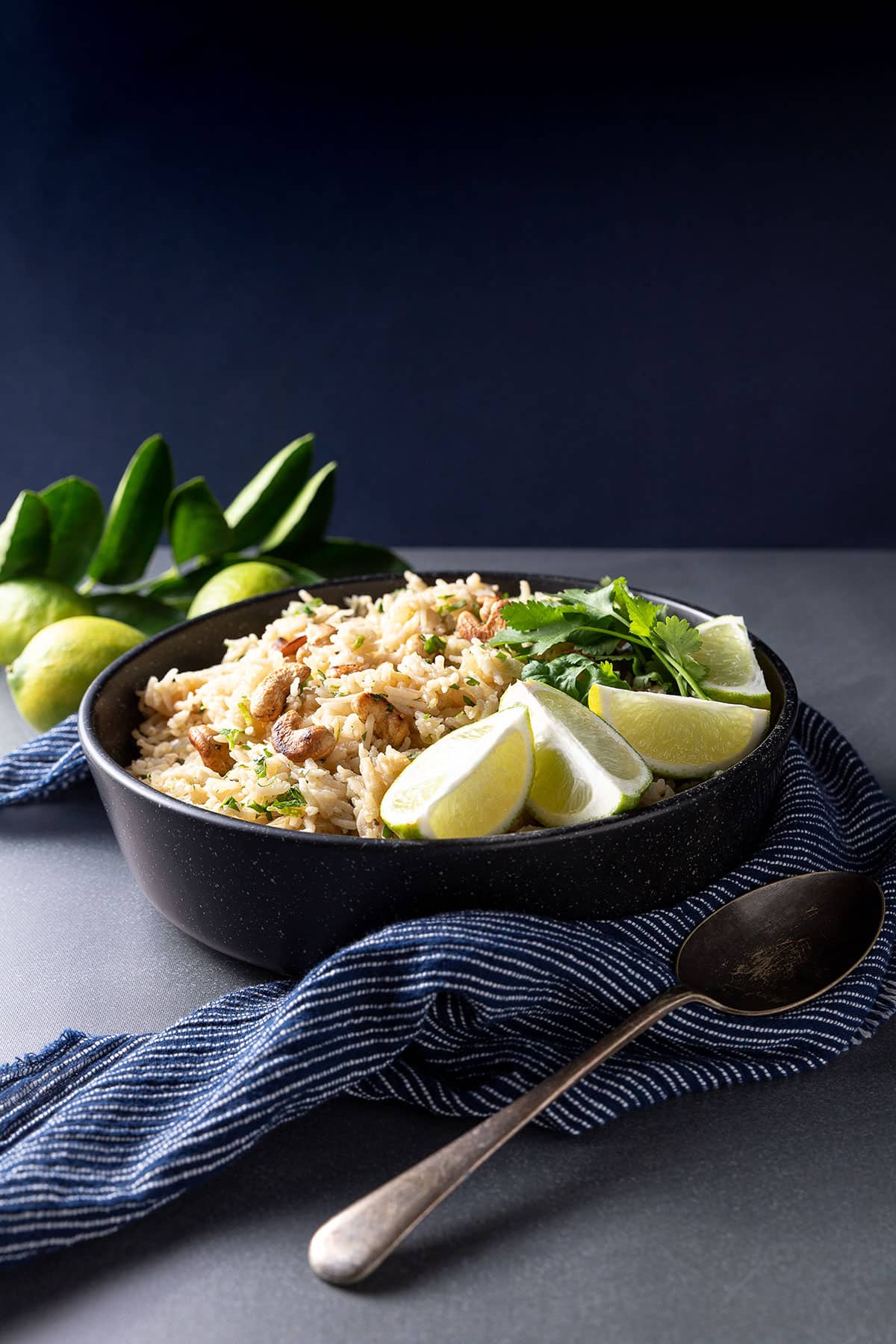 A black bowl containing rice pilaf, with cashew nuts, coriander (cilantro) and lime segments visible. 