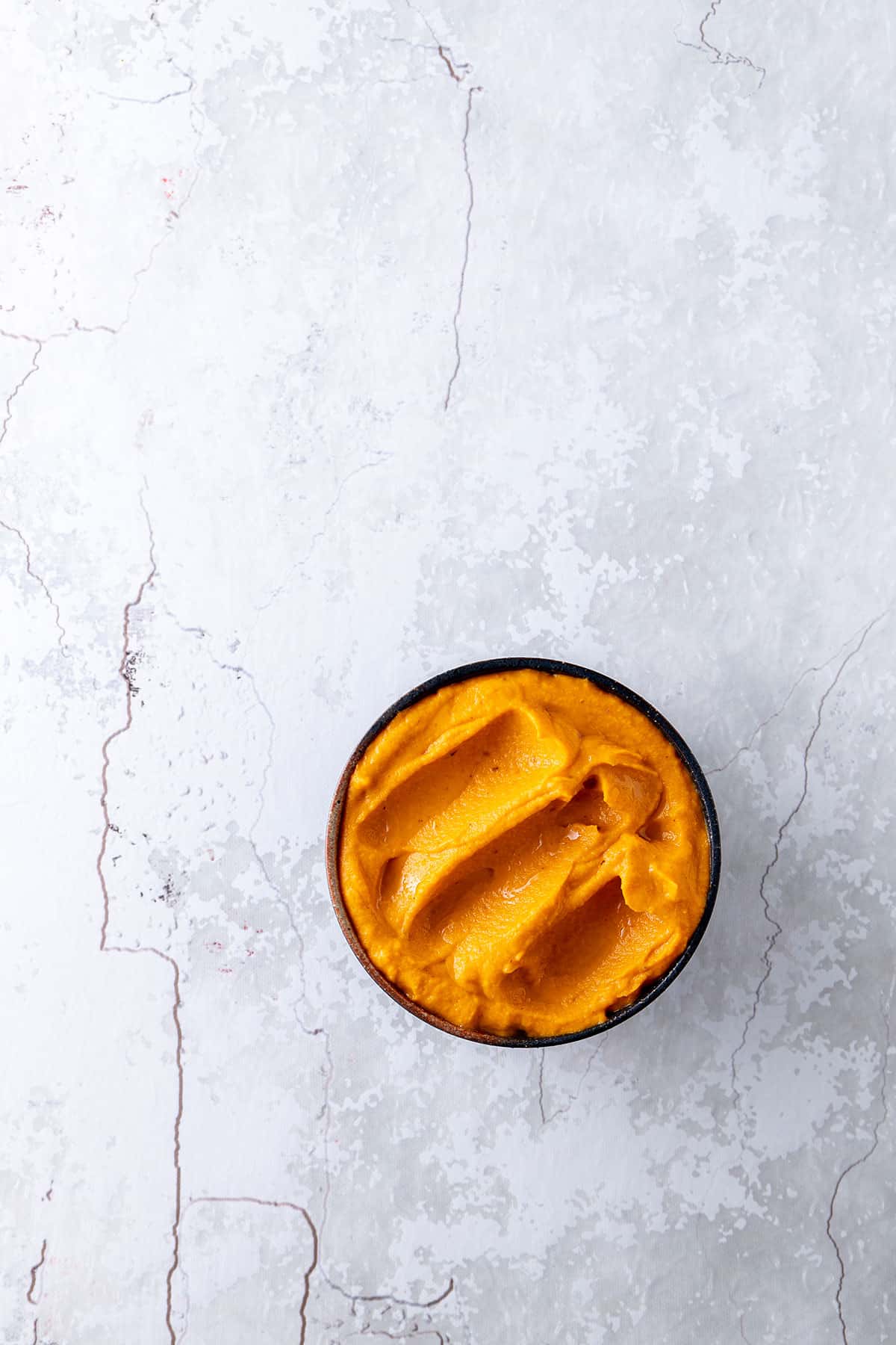 A bowl of bright orange carrot and harissa dip, sitting on a pale grey stone benchtop. 