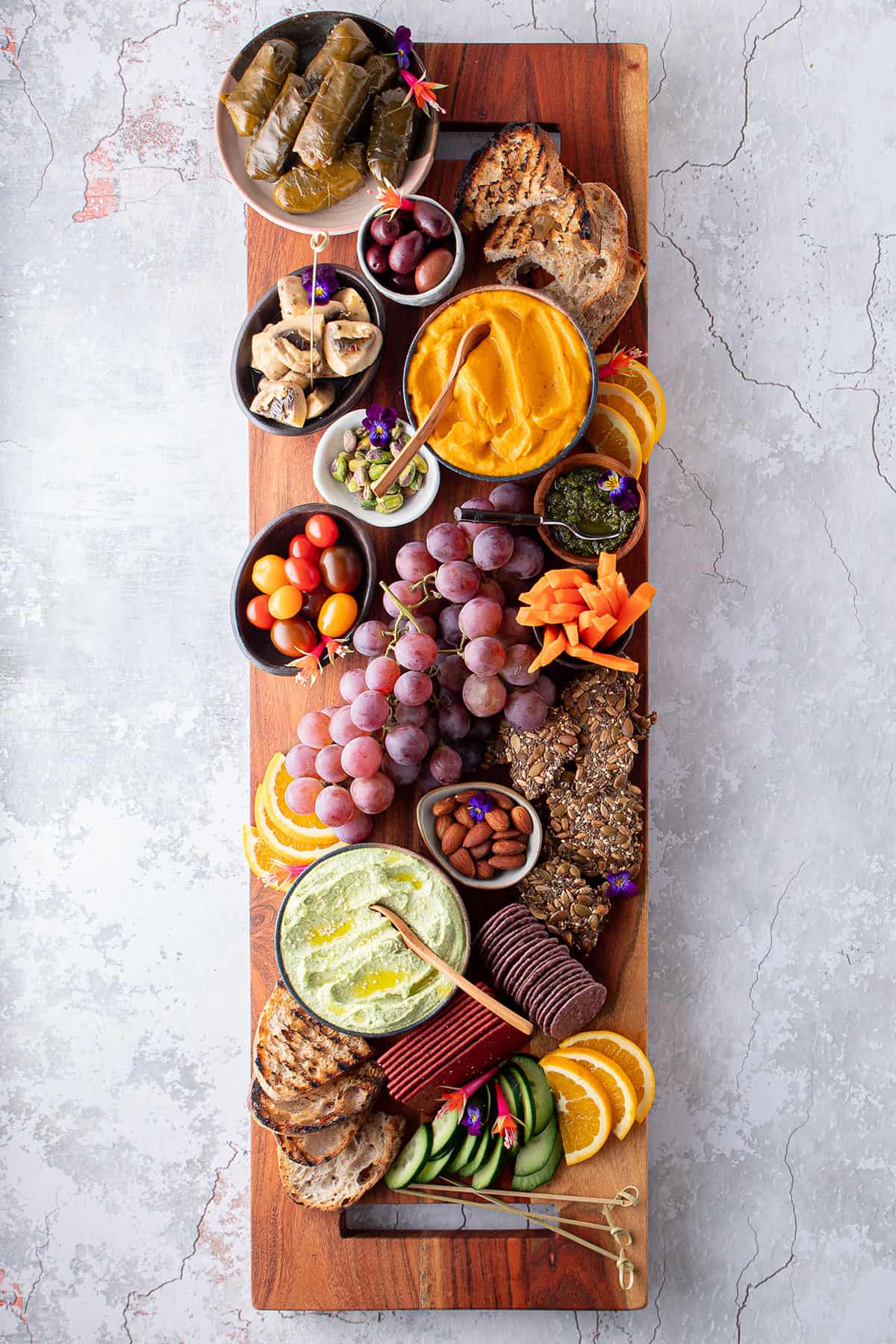 A wooden board with a variety of crackers, sliced carrot, orange, cucumber, grapes, nuts and dips. 