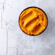 A bowl of bright orange carrot and harissa dip, sitting on a pale grey stone benchtop.
