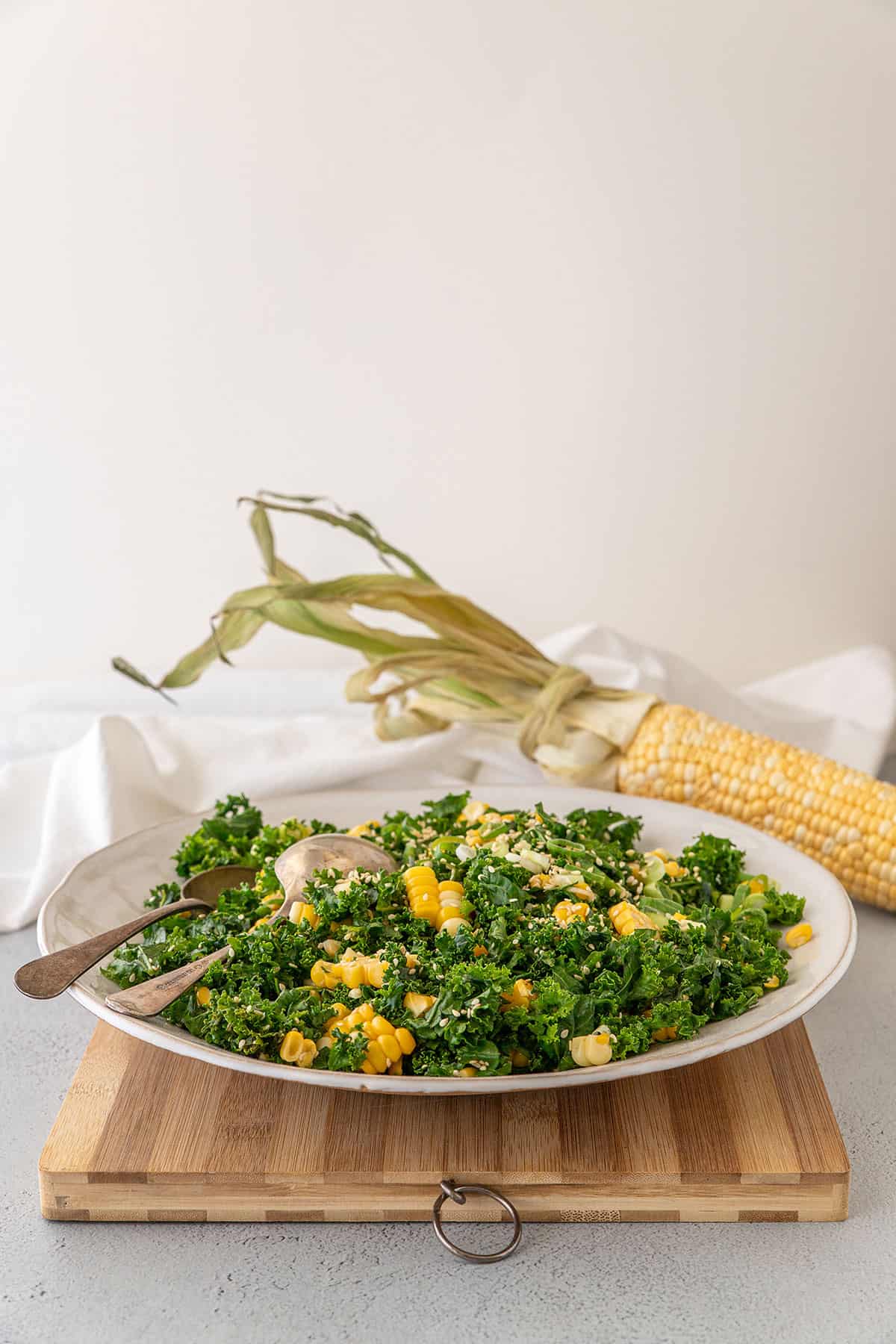 A wide white bowl with sweetcorn and kale salad in it, sitting on a bamboo chopping board. A whole cob of corn is in the background.