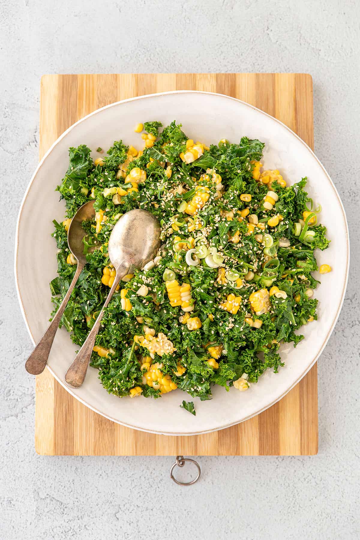 A wide white bowl with sweetcorn and kale salad in it, sitting on a bamboo chopping board. A whole cob of corn is in the background. Photo taken from above.