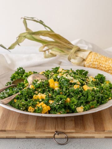A wide white bowl with sweetcorn and kale salad in it, sitting on a bamboo chopping board. A whole cob of corn is in the background.